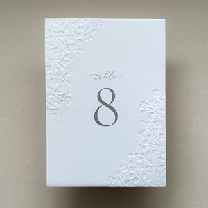May include: A rectangular white table number card featuring the text "table 8" in a grey serif font. The card is decorated with embossed floral patterns along the top-left and bottom-right corners, creating a textured, raised design on the paper.
