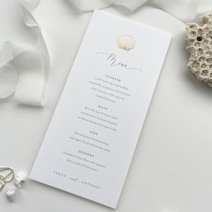 May include: A white wedding menu card with gold foil accents. The menu features a starter, main course, side dish, and dessert. The card is decorated with a gold seashell illustration and the text "Menu" in elegant script.