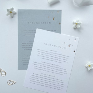 May include: Two light blue and white cards with gold lettering provide information about a wedding venue. The cards have the words "Information" and "Directions" printed on them. The cards also have a crescent moon and stars printed on them.