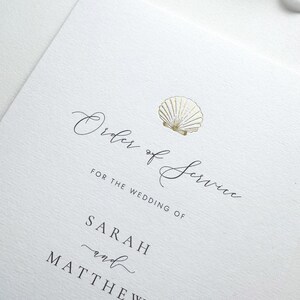 May include: A white wedding order of service card with elegant script. The text reads "Order of Service" and "For the Wedding of Sarah and Matthew." A gold seashell graphic is at the top. The paper has a textured finish.