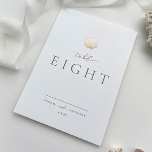 May include: White card with a gold seashell design and the text "table EIGHT" in black. Below the text is a line with the text "SARAH and ANTHONY 6.8.25" in black.