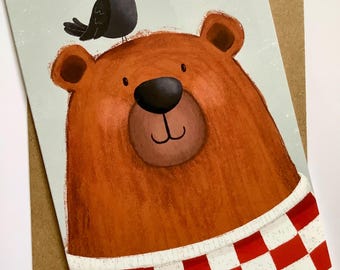 Puede incluir: Tarjeta de felicitación con una ilustración de un oso pardo y un pájaro negro. El oso lleva una bufanda a cuadros rojos y blancos. La tarjeta está sobre un sobre marrón, ideal para enviar un mensaje amistoso.