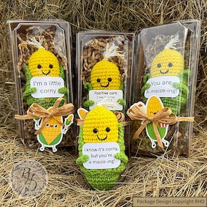 Könnte beinhalten: Handgefertigte gehäkelte Mais-Plüschtiere in transparenten Boxen mit braunem Papier. Jedes Plüschtier hat ein lächelndes Gesicht und hält ein Schild mit einem Wortspiel. Auf einem steht "I'm a little corny", auf einem anderen "Positive corn" und auf einem dritten "You are A-maize-ing!"