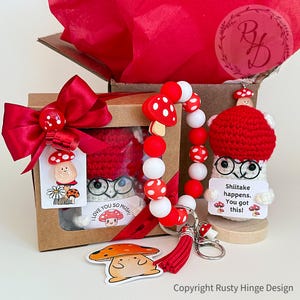 A boxed crochet mushroom gift set featuring a handmade crochet plush wearing cute glasses, colorful red and white beaded keychain, sticker, resin charm and gift tags with the pun 'Shiitake happens. You got this!’ Customizable gift box loaded with mushroom charm. Perfect emotional support, birthday, Coworker, Christmas or cheering-up gift.