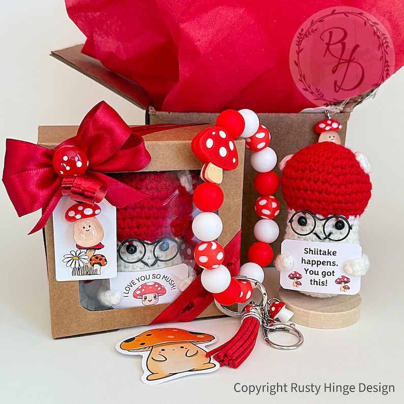 A boxed crochet mushroom gift set featuring a handmade crochet plush wearing cute glasses, colorful red and white beaded keychain, sticker, resin charm and gift tags with the pun 'Shiitake happens. You got this!’ Customizable gift box loaded with mushroom charm. Perfect emotional support, birthday, Coworker, Christmas or cheering-up gift.
