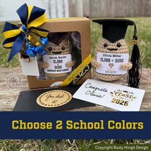 May include: Two graduation themed crocheted peanut figures, one in a box with a yellow and blue bow, and the other with a sign that says "Congrats Olivia!" and a sticker that says "Class of 2025".