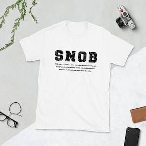 May include: White t-shirt with a black graphic that says "SNOB" and defines the word as a person who judges the importance of people mainly by their social position or wealth.