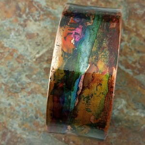 May include: A copper cuff bracelet with a colorful abstract design. The design features shades of blue, green, orange, and pink.
