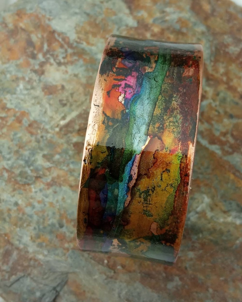 May include: A copper cuff bracelet with a colorful abstract design. The design features shades of blue, green, orange, and pink.