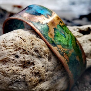 May include: A wide copper cuff bracelet with a colorful patina of blues, greens, and browns. The bracelet has a curved shape and is displayed on a piece of driftwood. The colors create a unique, artistic look.