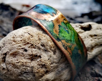 May include: A wide copper cuff bracelet with a colorful patina of blues, greens, and browns. The bracelet has a curved shape and is displayed on a piece of driftwood. The colors create a unique, artistic look.