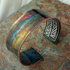May include: A copper cuff bracelet with a colorful abstract design. The inside of the bracelet has a black and white sticker with the text "Cornish Kissed".