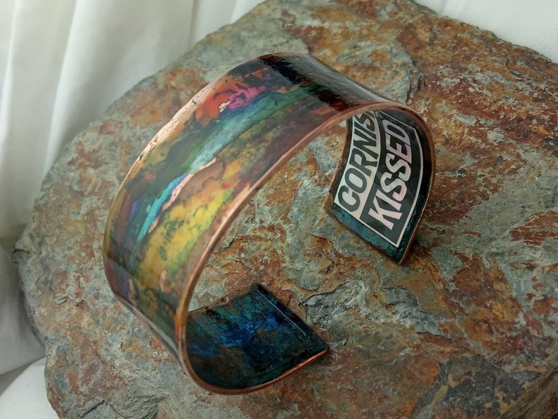 May include: A copper cuff bracelet with a colorful abstract design. The inside of the bracelet has a black and white sticker with the text "Cornish Kissed".