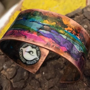 May include: A copper bracelet featuring a colourful abstract design and the words "Inspired by the Sea, Ocean-Kissed" on the side.