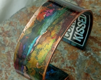 May include: A copper cuff bracelet with a colorful, abstract design. The design features shades of blue, green, yellow, orange, and pink. The bracelet is slightly curved and has a smooth, polished finish.