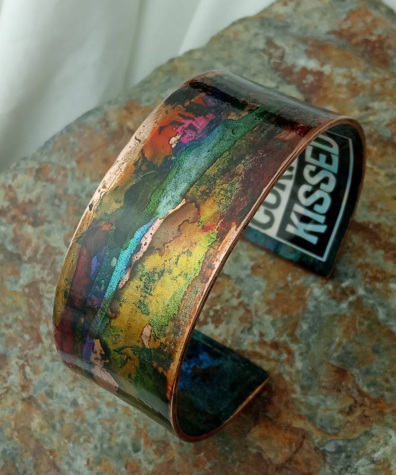 May include: A copper cuff bracelet with a colorful, abstract design. The design features shades of blue, green, yellow, orange, and pink. The bracelet is slightly curved and has a smooth, polished finish.