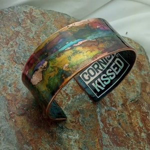 May include: A copper bracelet with a colorful abstract design and the words "CORN KISSED" on the inside.