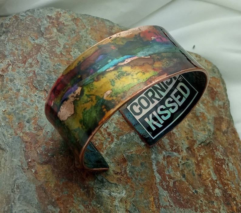 May include: A copper bracelet with a colorful abstract design and the words "CORN KISSED" on the inside.