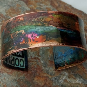 May include: A wide copper bracelet with a colorful, abstract design. The design features a band of green, blue, and pink hues, with hints of orange and brown. The bracelet is slightly curved and has a smooth, polished finish.