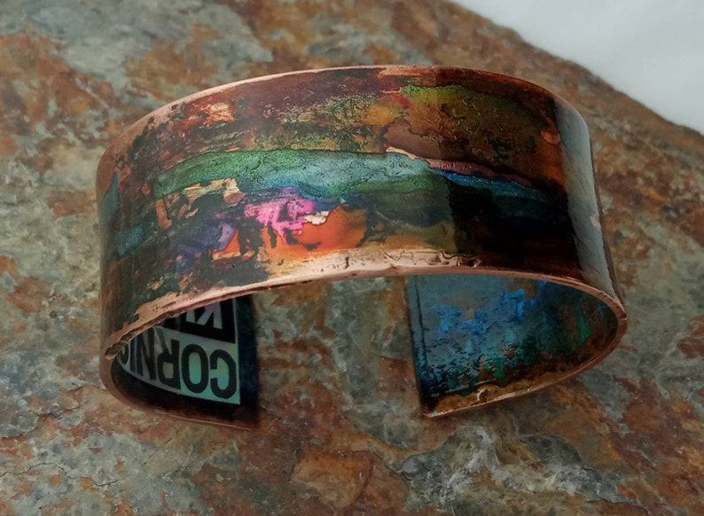 May include: A wide copper bracelet with a colorful, abstract design. The design features a band of green, blue, and pink hues, with hints of orange and brown. The bracelet is slightly curved and has a smooth, polished finish.