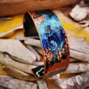 May include: A copper bracelet with a blue and green patina. The bracelet is wide and has a rounded shape. The patina is uneven and has a mottled appearance.