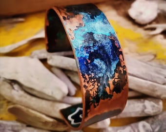 May include: A copper bracelet with a blue and green patina. The bracelet is wide and has a rounded shape. The patina is uneven and has a mottled appearance.