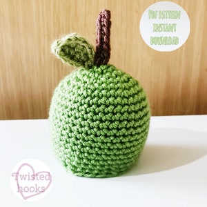 May include: A handmade green crochet apple with a brown stem and a green leaf. The apple is round and textured, with the text "PDF PATTERN INSTANT DOWNLOAD" in a white circle. The logo "Twisted hooks" is in a pink heart.
