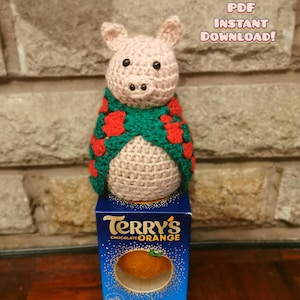 Peut inclure: Une figurine de cochon crochetée, vêtue d'une écharpe verte et rouge, posée sur une boîte de Terry's Chocolate Orange. Le cochon est rose clair avec des yeux en boutons noirs. La boîte est bleue avec les mots "Terry's Chocolate Orange". Le texte de l'image indique "PDF TÉLÉCHARGEMENT INSTANTANÉ!"
