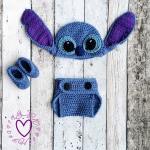 May include: A handmade crochet baby set in blue, featuring a hat with large ears, a diaper cover, and a pair of booties. The hat has large eyes and the ears are purple. The set is designed to resemble a cartoon character.