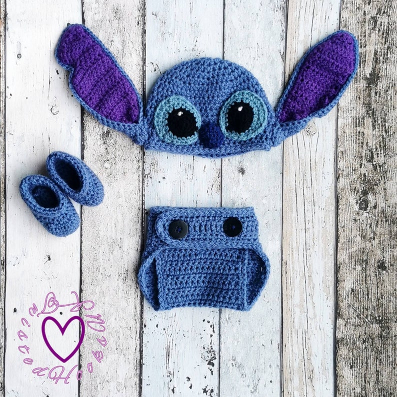 May include: A handmade crochet baby set in blue, featuring a hat with large ears, a diaper cover, and a pair of booties. The hat has large eyes and the ears are purple. The set is designed to resemble a cartoon character.