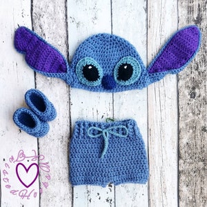 May include: A blue crochet baby outfit, including a hat with large ears, shorts, and booties. The hat has large black and white eyes and a small blue nose. The outfit is displayed on a light wood-grain surface.