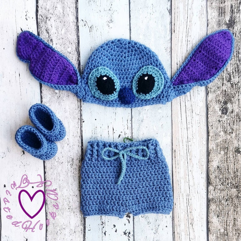May include: A blue crochet baby outfit, including a hat with large ears, shorts, and booties. The hat has large black and white eyes and a small blue nose. The outfit is displayed on a light wood-grain surface.