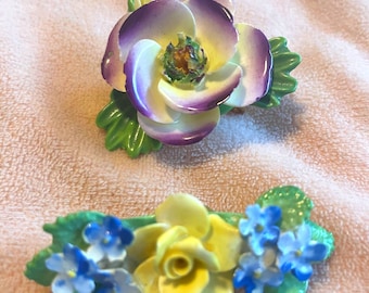 Puede incluir: Dos broches de porcelana con forma de flores. El broche superior presenta una flor grande de color morado y blanco con hojas verdes. El broche inferior presenta una rosa amarilla con flores azules y hojas verdes.