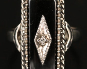 May include: A silver ring with a black onyx stone set in a rectangular frame. The frame is decorated with a rope-like design and features a small diamond in the center of the onyx stone.