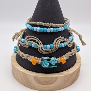 May include: Three handmade bracelets displayed on a black cone-shaped stand. The bracelets feature turquoise and blue beads, jute rope, and a decorative glass bead with a floral design. The stand sits on a wooden base.