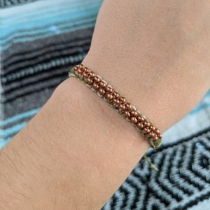 Op de afbeelding: Een groene en witte vriendschapsband met koperen kralen. De armband is om een pols gebonden.