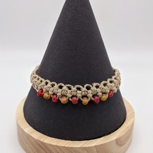 Puede incluir: Un brazalete de macramé beige con cuentas rojas y ámbar. El brazalete se muestra en un soporte cónico negro con una base de madera.