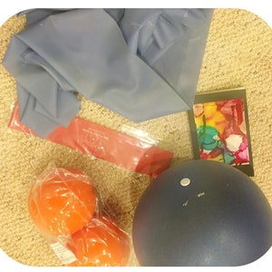 Puede incluir: Una banda de ejercicio azul, una banda de resistencia roja, dos pelotas de ejercicio naranjas, una pelota de ejercicio azul y una tarjeta pequeña con un diseño abstracto colorido.
