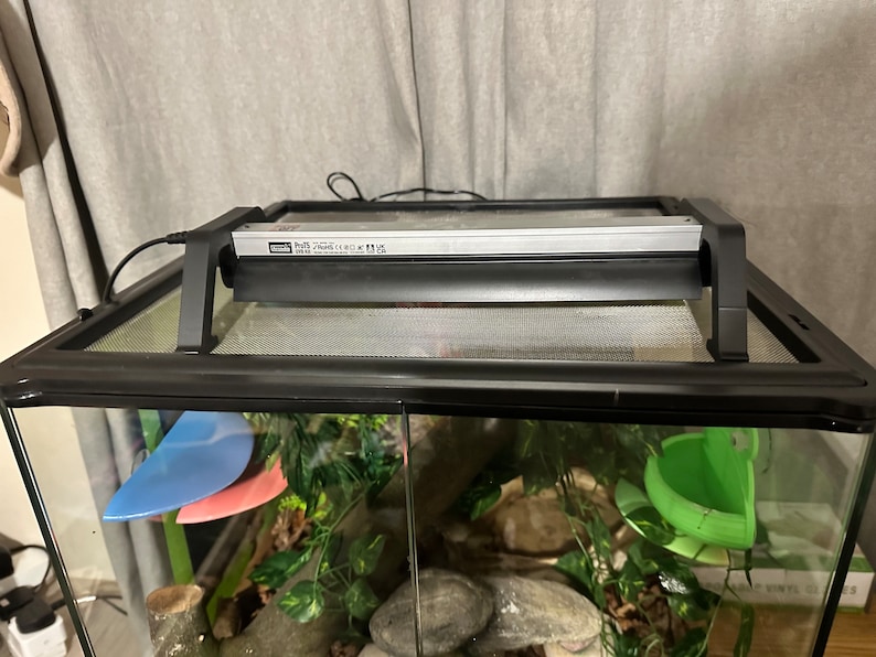 Puede incluir: Terrario negro para reptiles con una tapa de malla y una l&aacute;mpara de calor Zoo Med Repti Basking Spot. El terrario tiene una planta verde frondosa, una rama marr&oacute;n y un bebedero de pl&aacute;stico verde.