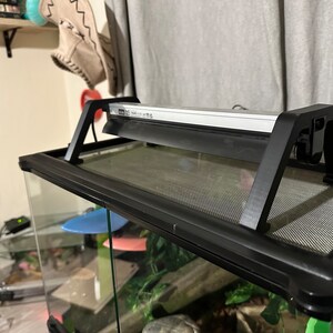 Puede incluir: Un terrario negro para reptiles con una tapa de malla y un accesorio de luz negro fijado en la parte superior. El accesorio de luz tiene una barra de luz blanca y un cable negro.