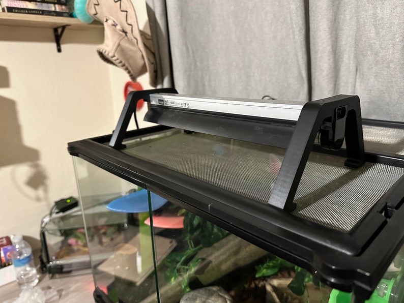 Puede incluir: Un terrario negro para reptiles con una tapa de malla y un accesorio de luz negro fijado en la parte superior. El accesorio de luz tiene una barra de luz blanca y un cable negro.