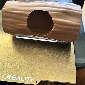 Puede incluir: Un objeto marrón en forma de tronco con una gran abertura circular. El objeto está sobre una superficie dorada con la palabra "CREALITY" impresa. El tronco parece ser un artículo decorativo o funcional.