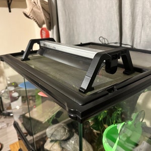 Puede incluir: Terrario negro para reptiles con una tapa de malla y un accesorio de luz negro fijado en la parte superior. El terrario tiene un bebedero verde y una rueda de pl&aacute;stico blanca en el interior.