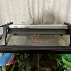 May include: A black reptile terrarium with a black metal mesh top. A silver heat lamp with a black reflector is mounted on the top of the terrarium. The lamp has a label that reads "Zoo Med ReptiSun 5.0 UVB 10.0", and the cord is visible.
