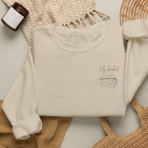 Könnte beinhalten: Ein cremefarbenes Sweatshirt mit Rundhalsausschnitt und einer kleinen, schwarzen Strichgrafik auf der linken Brust. Das Design zeigt einen Stapel aus drei Büchern mit einem aufgeschlagenen Buch darauf und dem handschriftlichen Text "fully booked".