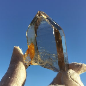 May include: A large, clear quartz crystal with a golden brown inclusion. The crystal is being held by two gloved hands against a bright blue sky.
