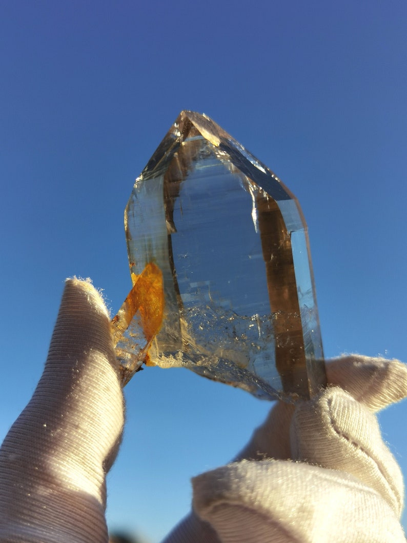May include: A large, clear quartz crystal with a golden brown inclusion. The crystal is being held by two gloved hands against a bright blue sky.