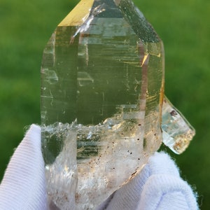 May include: A large, clear quartz crystal with a pointed tip and a natural, rough texture. The crystal has a slight yellow hue and is held in a white glove.