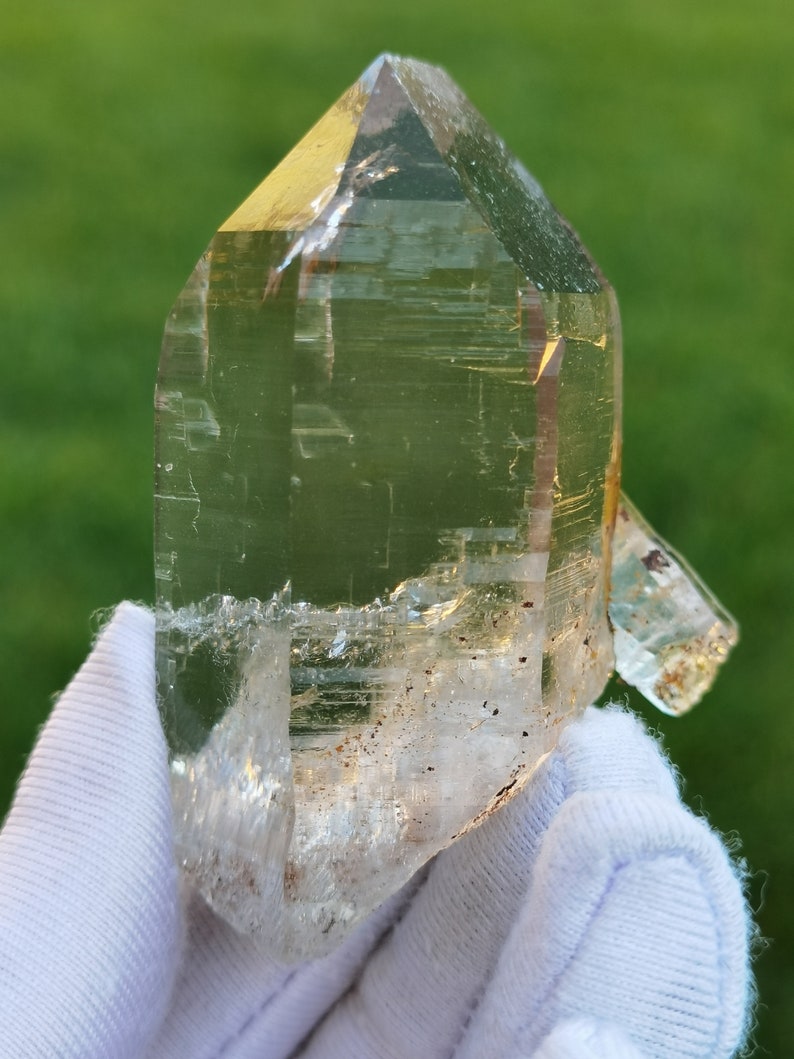 May include: A large, clear quartz crystal with a pointed tip and a natural, rough texture. The crystal has a slight yellow hue and is held in a white glove.