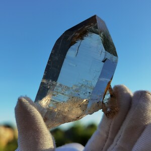 May include: A large, clear quartz crystal with a point, held in gloved hands. The crystal has a slight brown tint and is translucent.